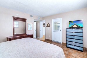 A bedroom with a large bed, a dresser, a television, and a door leading to another room.