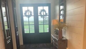 A hallway with wooden flooring, a dark door with a wreath, and a small table with a lamp and a vase. - Modern farmhouse with a pool (Cat Spring)