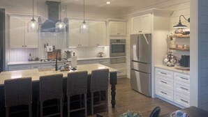 A modern kitchen with white cabinets, a central island with bar stools, stainless steel appliances, pendant lighting, and a built-in oven. - Modern farmhouse with a pool (Cat Spring)