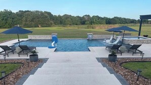 A swimming pool with two slides, surrounded by lounge chairs and umbrellas, with a view of a golf course in the background. - Modern farmhouse with a pool (Cat Spring)