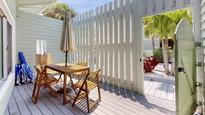 En overdækket terrasse med et træbord og stole, et hvidt stakværkshegn og en dør, der fører til en have.