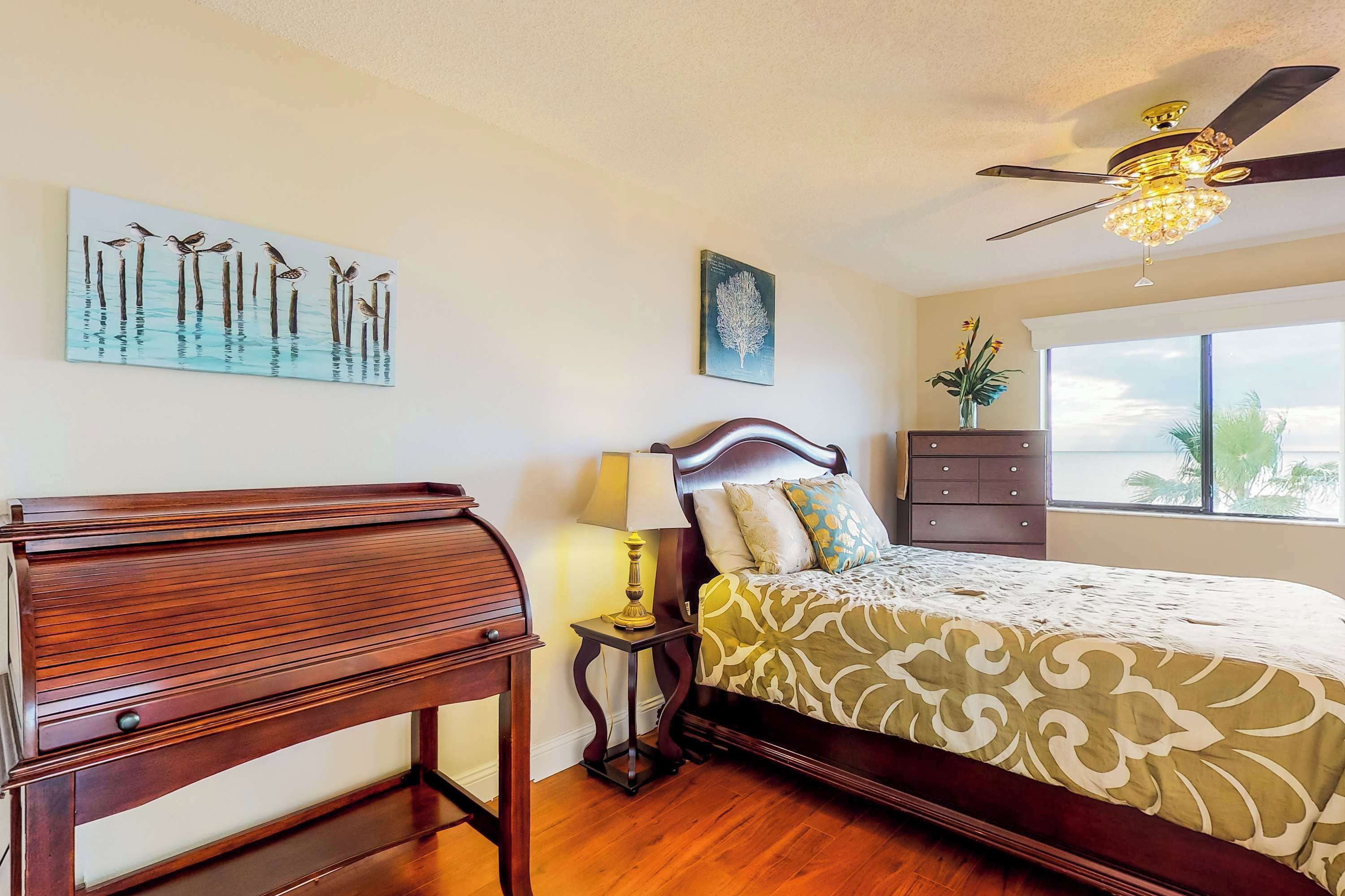 A bedroom with a wooden bed, a ceiling fan, a nightstand with a lamp, and artwork on the walls.