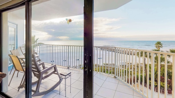 A balcony with a view of the ocean, a white railing, and a table with chairs.