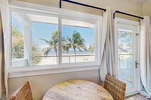 A room with a large window overlooking palm trees and a beach. There is a round table with two wicker chairs. - Gulf front condo top floor beach access full kitchen and central AC (Indian Shores)