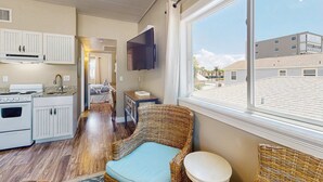 A compact kitchen with a stove, cabinets, and a television. A wicker chair is near a window with a view of buildings. - Gulf front condo top floor beach access full kitchen and central AC (Indian Shores)