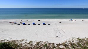 En strand med hvidt sand, klart blåt vand og enkelte personer. Blå strandstole og parasoller er placeret der.