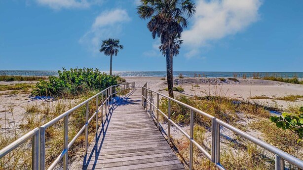 Una pasarela de madera que lleva a una playa con palmeras y un cielo azul despejado.