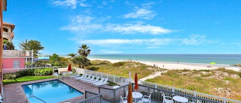 A pool area with lounge chairs and tables, a view of the beach, and a clear sky.