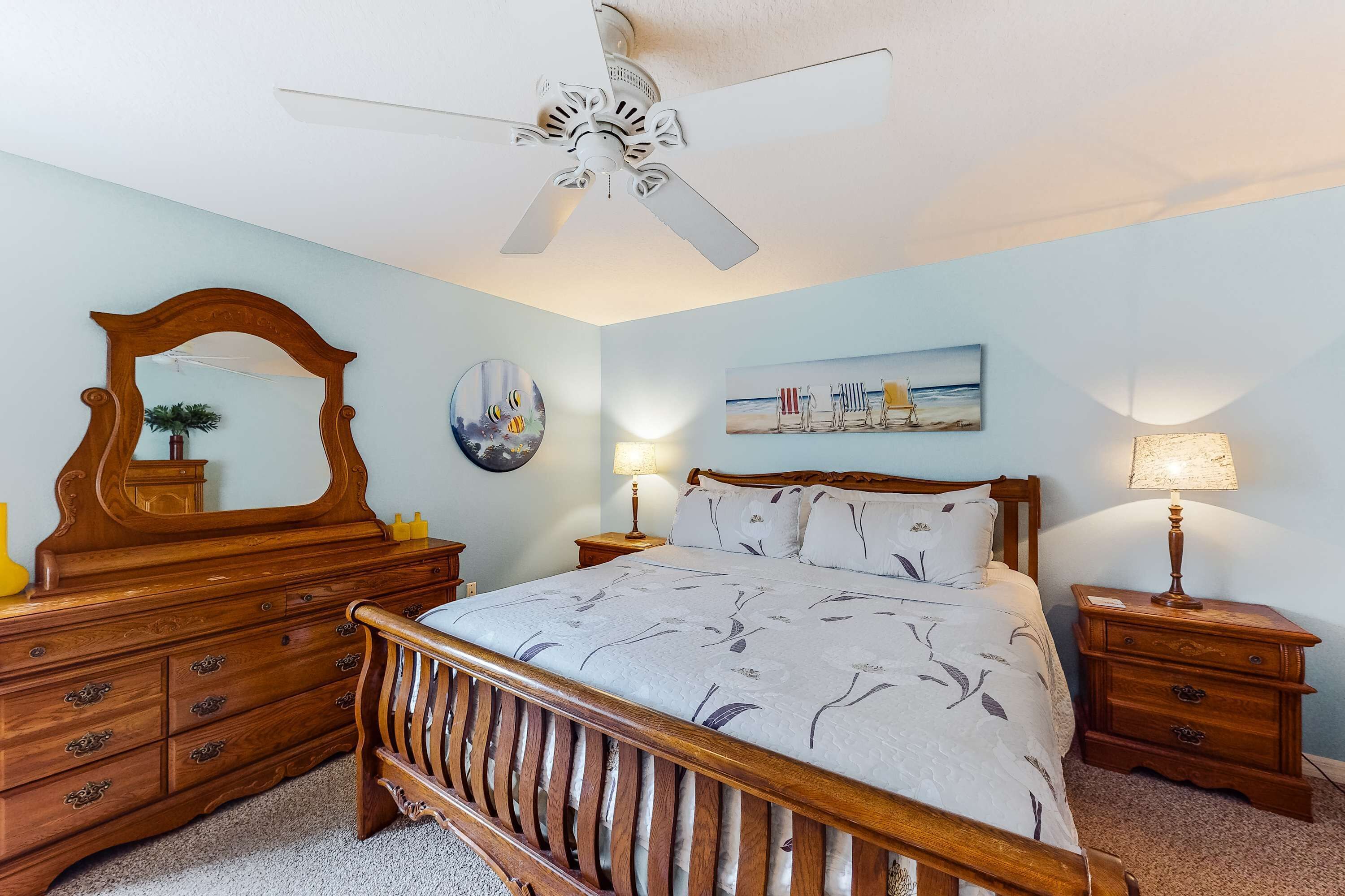 A bedroom with a wooden bed, a ceiling fan, a dresser, a nightstand, a lamp, a mirror, and a painting on the wall.