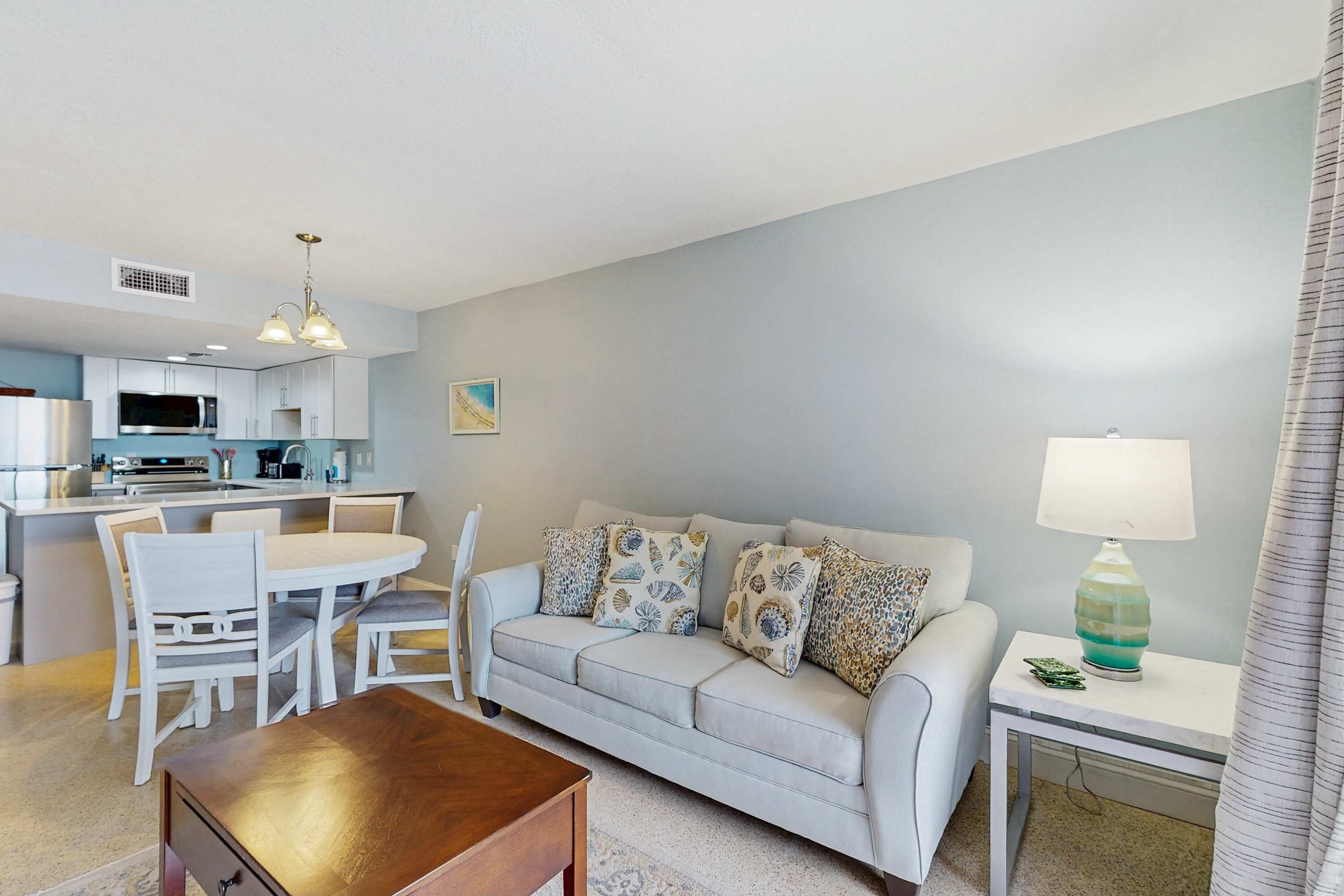 A modern living room with a sofa, dining table, and chairs. There is a kitchen area with cabinets and appliances. A lamp is on a side table.