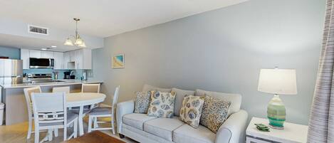 A modern living room with a sofa, dining table, and chairs. There is a kitchen area with cabinets and appliances. A lamp is on a side table.