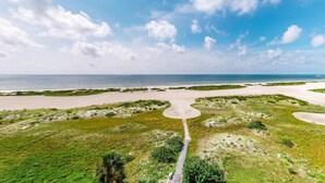 An aerial view of a coastal landscape with a sandy beach, grassy areas, and a clear sky.