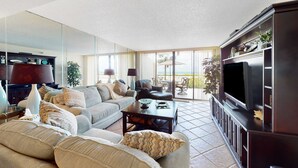 A living room with a sofa, coffee table, and TV stand. There is a large window and a door leading to an outdoor patio.
