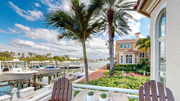 A balcony with wooden chairs, a table with two potted plants, a view of a marina with boats, and a building with arched windows. - Dog Friendly Retreat w/Bay Views, Pool, Spa & Onsite Marina (Clearwater)