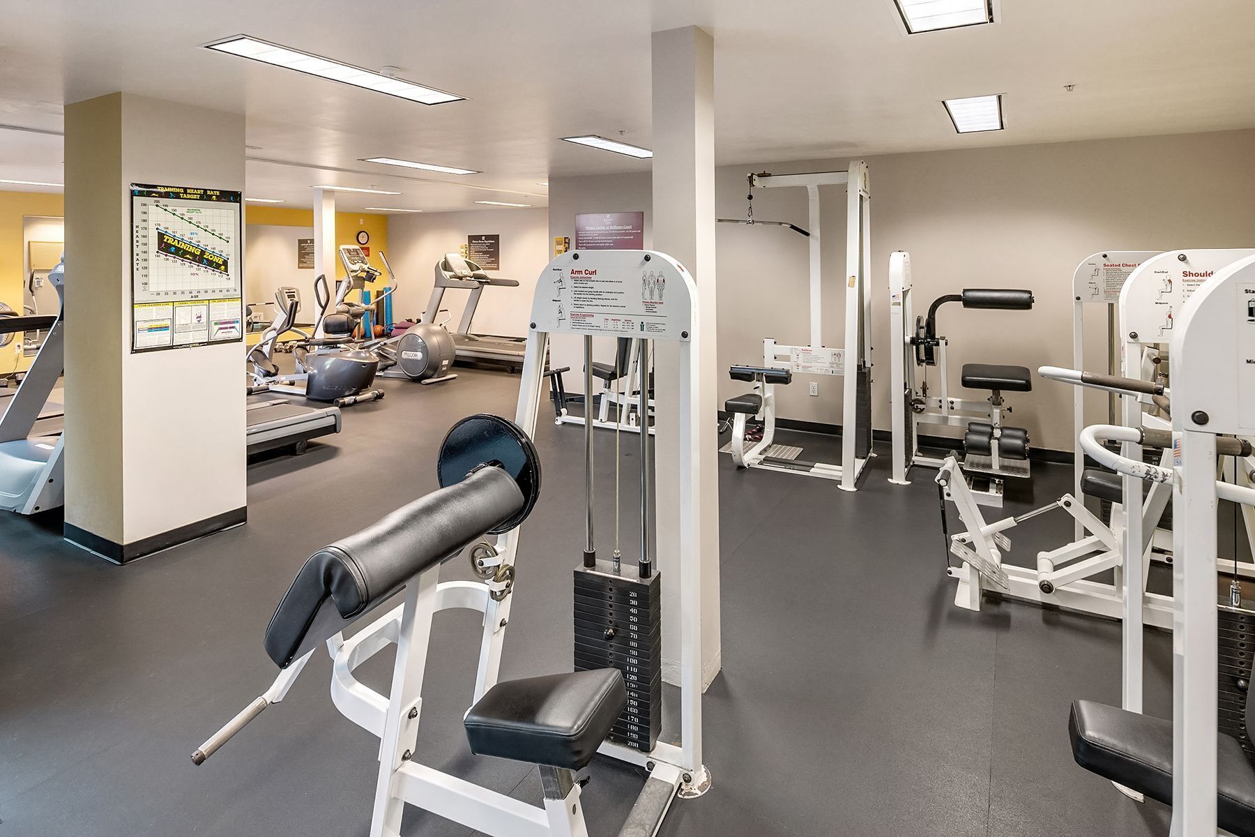 A gym with various exercise machines, a treadmill, and a wall with instructional posters.
