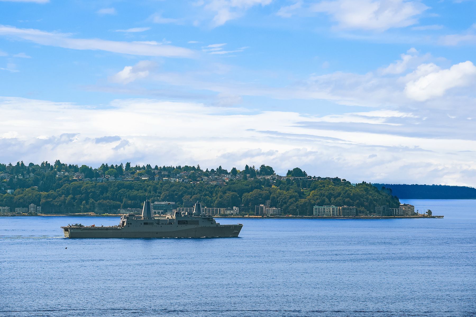 Un navire militaire sur une étendue d’eau calme, avec un paysage vallonné en arrière-plan.