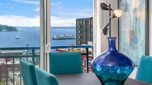 Una stanza con un'ampia finestra che si affaccia su un corpo d'acqua, una nave e un edificio. Si nota un tavolo con un vaso blu e delle sedie.
