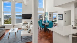 Un'ampia zona giorno con una grande finestra vista su uno specchio d'acqua, un'area pranzo con sedie color petrolio e una cucina dal design moderno.