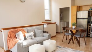 Ein modernes Wohnzimmer mit einem grauen Sofa, Poufs, einem Couchtisch und einer Lampe. Im Hintergrund ist eine Küchenzeile mit Holzschränken und einem Kühlschrank zu sehen.