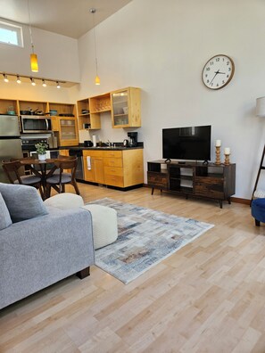 A modern kitchen with wooden cabinets, a built-in microwave, and a coffee maker. There is a dining area with a table and chairs, and a living room with a sofa and a TV. - Townhome • Hospital District • King Bed + Garage (Billings)