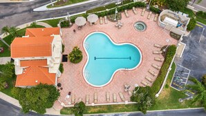 Piscina con sillas de descanso y sombrillas, rodeada de un jardín ajardinado con palmeras y un edificio de techo naranja.