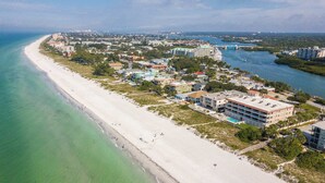 Luftaufnahme einer Küstenlandschaft mit einem langen Sandstrand, einem Yachthafen und einer Mischung aus Wohn- und Geschäftshäusern.