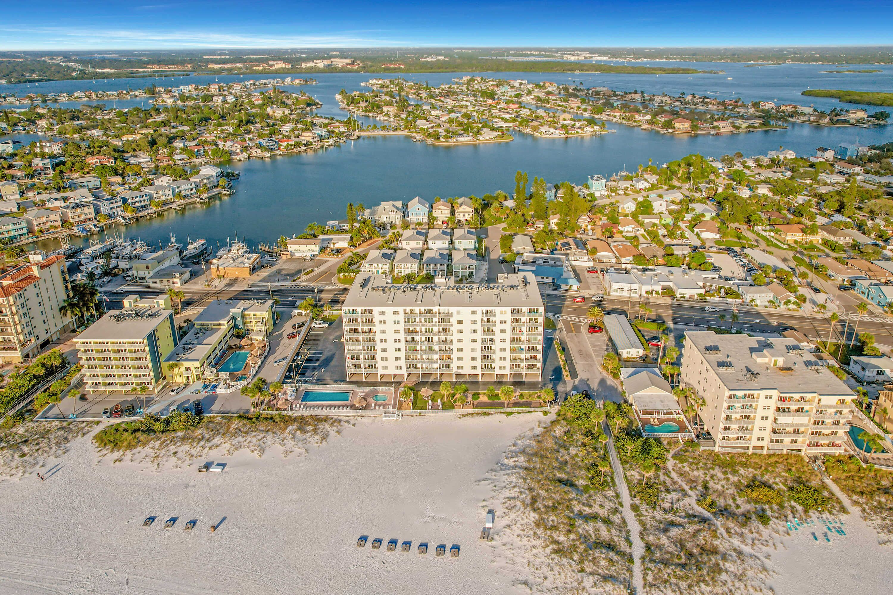 An aerial view of a coastal town with a prominent hotel featuring multiple floors, a beach with sun loungers, and a marina with boats.