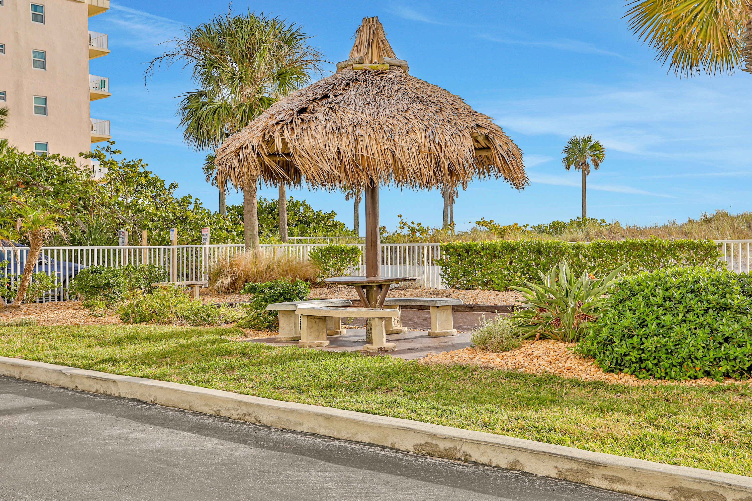 A thatched gazebo with benches, surrounded by well-maintained grass and palm trees, with a building in the background.