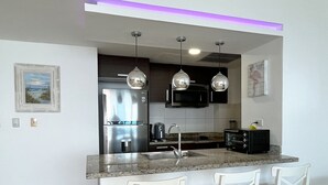 A modern kitchen with a stainless steel refrigerator, a microwave, and a toaster. There are three white bar stools and a granite countertop. - Charming 2-bedrooom Apartment with Majestic Ocean & Pool View in Juan Dolio (Juan Dolio)