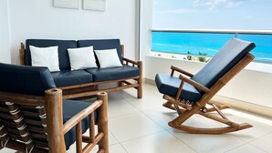 A balcony with a view of the ocean, a dark blue sofa with white cushions, and a wooden rocking chair. - Charming 2-bedrooom Apartment with Majestic Ocean & Pool View in Juan Dolio (Juan Dolio)