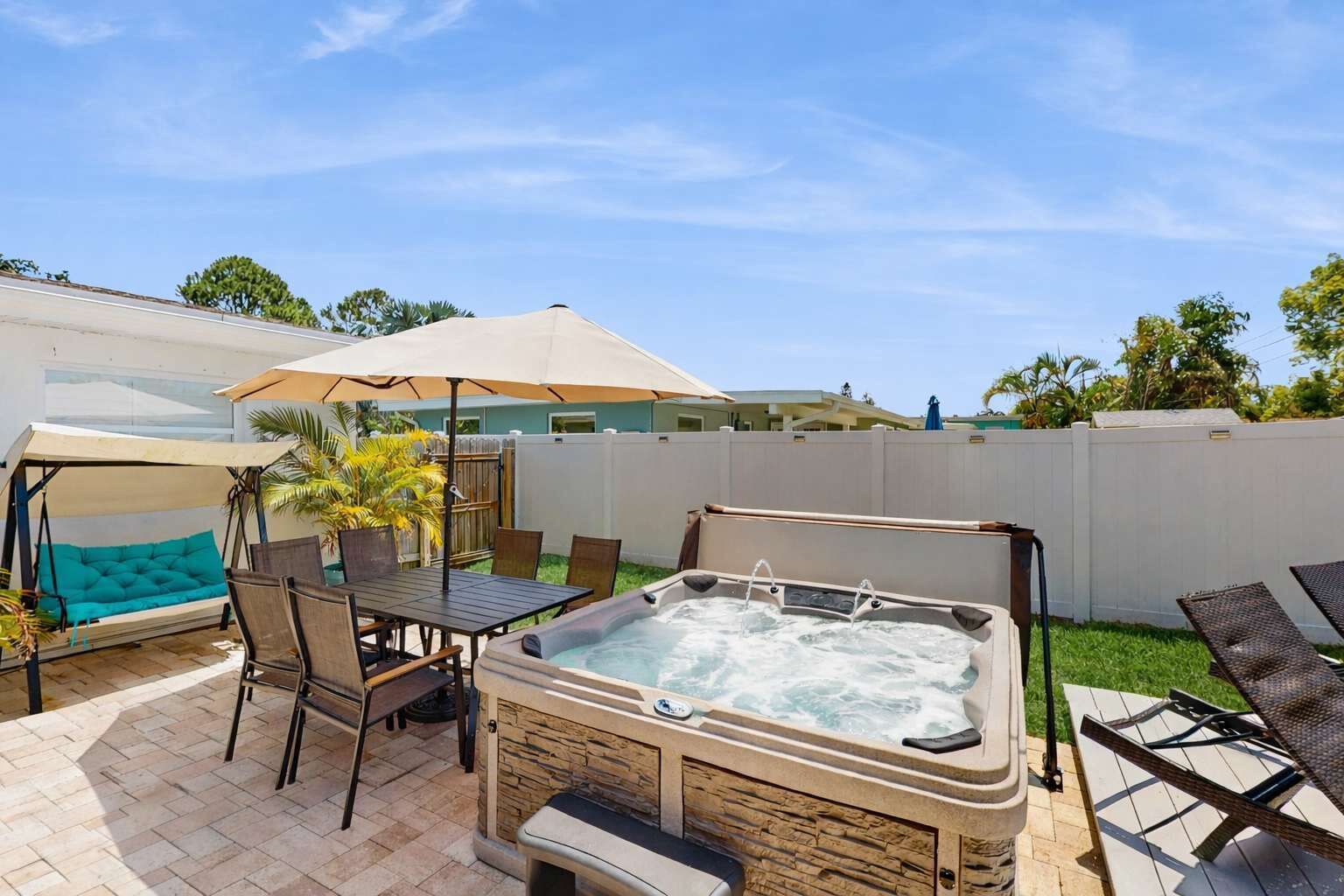 Un patio trasero con jacuzzi, una silla colgante, una mesa y un porche cubierto.