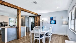 Une cuisine moderne dotée d’un îlot central, de poutres en bois et d’un coin repas avec des chaises blanches et une table en verre.