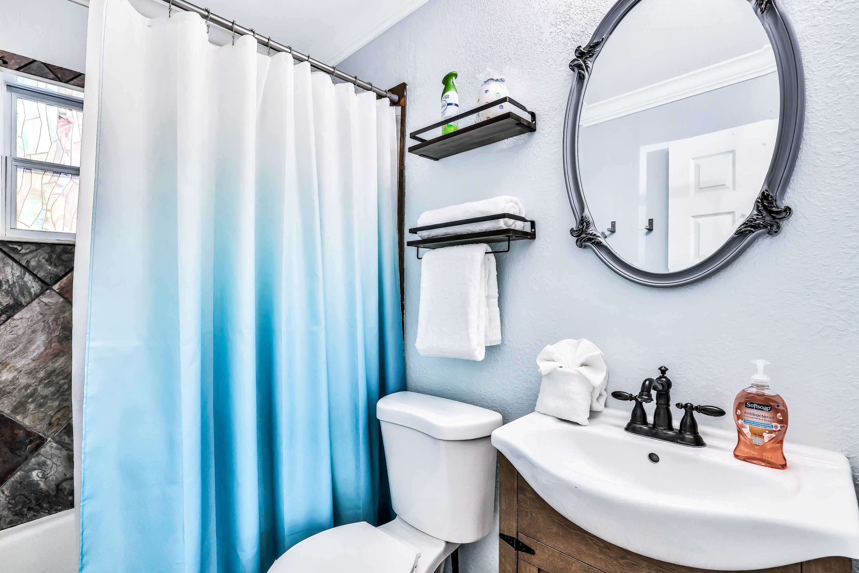 Une salle de bain avec un lavabo blanc, un miroir, un rideau de douche et des toilettes. Il y a deux étagères au-dessus du lavabo, l’une contenant une bouteille et l’autre une serviette.