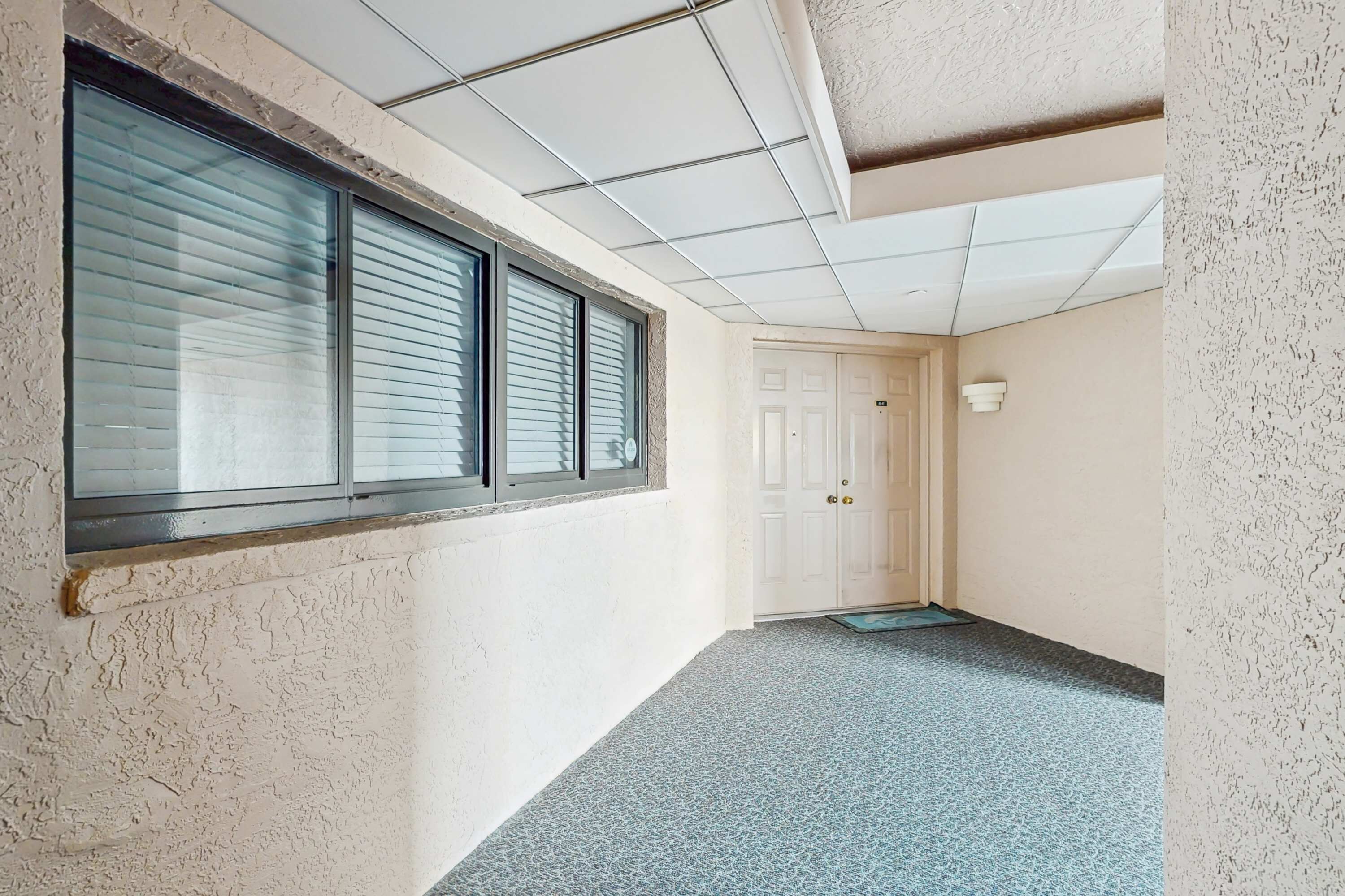 Een gang met een deur aan het einde, een raam met zonwering, en een structuurwand.