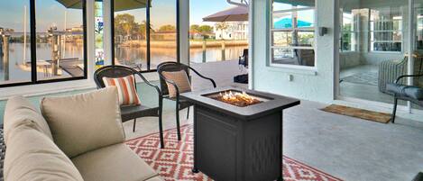 A covered patio with a fire pit, seating area, and large windows overlooking a body of water.
