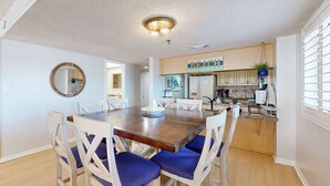 A dining area with a wooden table and white chairs with blue cushions, a kitchen with a 'BEACH' sign, and a round mirror on the wall. - Corner beach condo with Gulf views beachside Pool and Hot tub (Indian Shores)