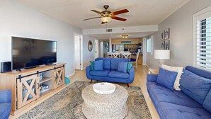 A living room with a blue sofa, a wooden entertainment unit, a ceiling fan, and a kitchen in the background. - Corner beach condo with Gulf views beachside Pool and Hot tub (Indian Shores)