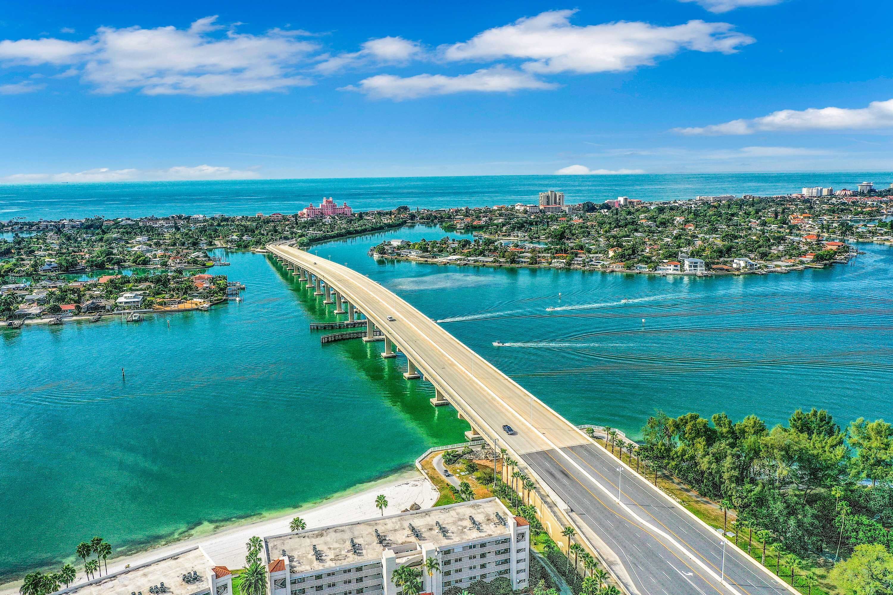 A coastal cityscape with a prominent bridge connecting two land masses, clear turquoise waters, and a mix of residential and commercial buildings.