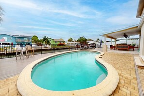 A swimming pool with a tiled deck, surrounded by a fence, and a covered patio area with outdoor furniture.