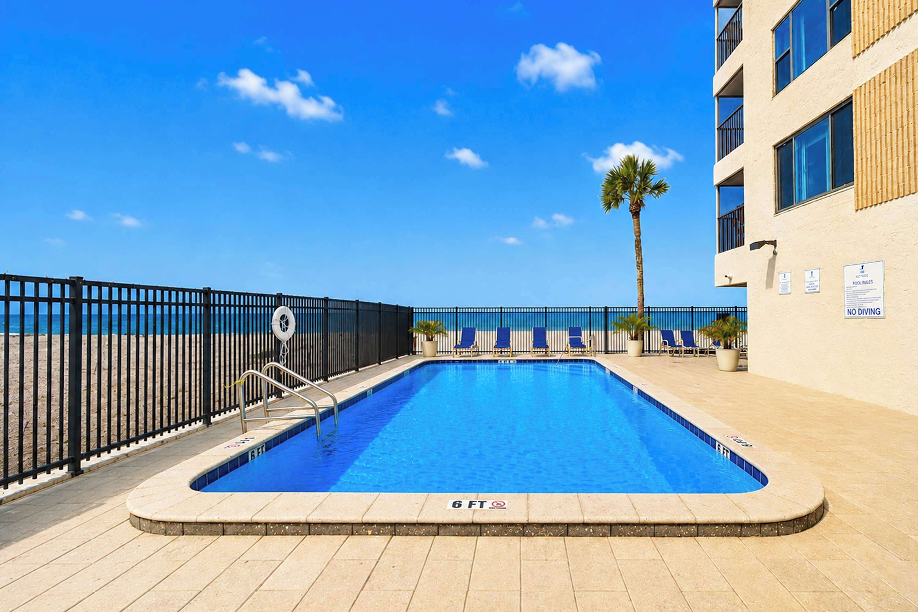 A swimming pool with a 6 ft depth, surrounded by a black fence, with a palm tree and lounge chairs in the background.