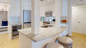 A modern kitchen with white cabinetry, a stainless steel refrigerator, a microwave, and a sink. There are two stools at the island counter. - Gulf front penthouse on Belleair Beach (Belleair Beach)