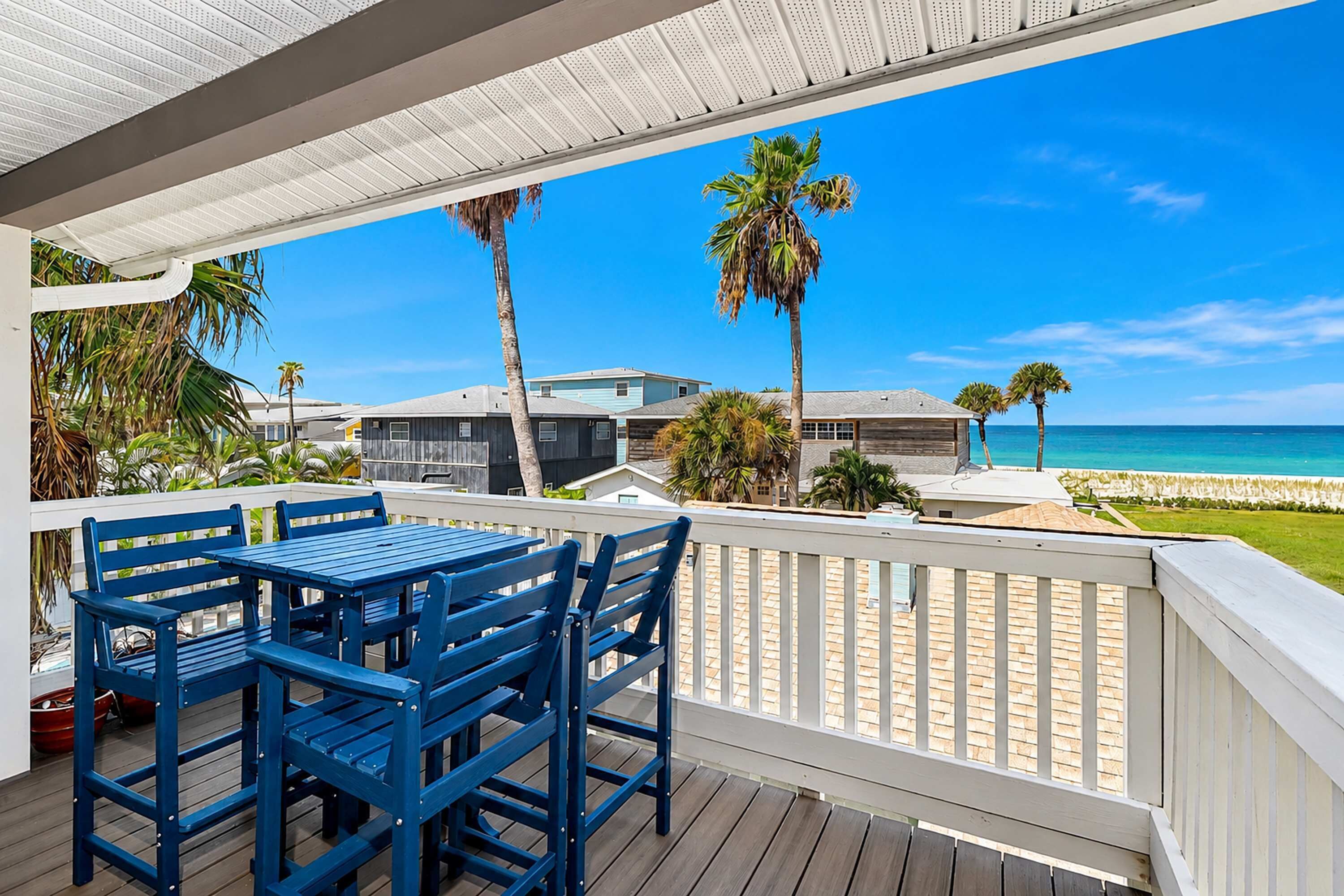 A wooden deck with blue chairs and a table, overlooking a beach with palm trees and a clear blue sky.