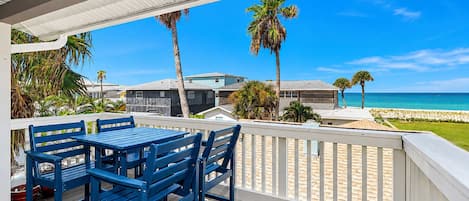A wooden deck with blue chairs and a table, overlooking a beach with palm trees and a clear blue sky.