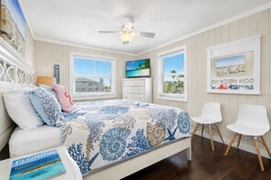 A bedroom with a bed, a ceiling fan, a TV, two chairs, and artwork on the walls. - Delightful condo with beach and pool access washer dryer (Belleair Beach)