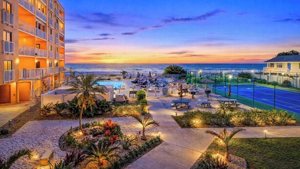 Un hotel con piscina, mobiliario exterior y vista al océano al atardecer.