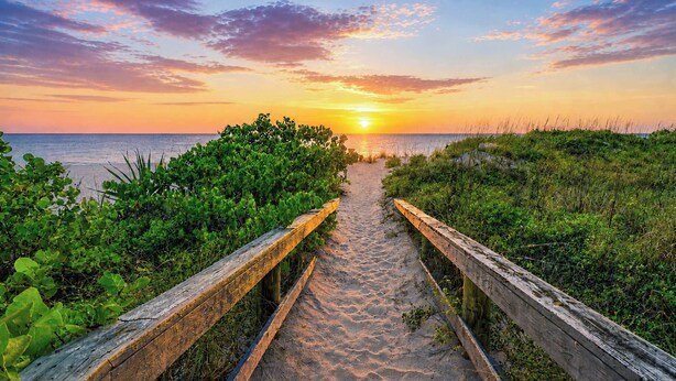 Ein hölzerner Weg führt zu einem Strand, im Hintergrund ist ein Sonnenuntergang zu sehen.