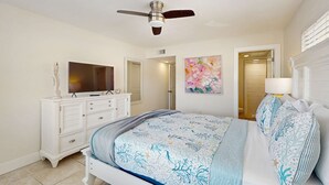 Une chambre à coucher avec un lit, un ventilateur de plafond, une télévision, une commode et un tableau accroché au mur.