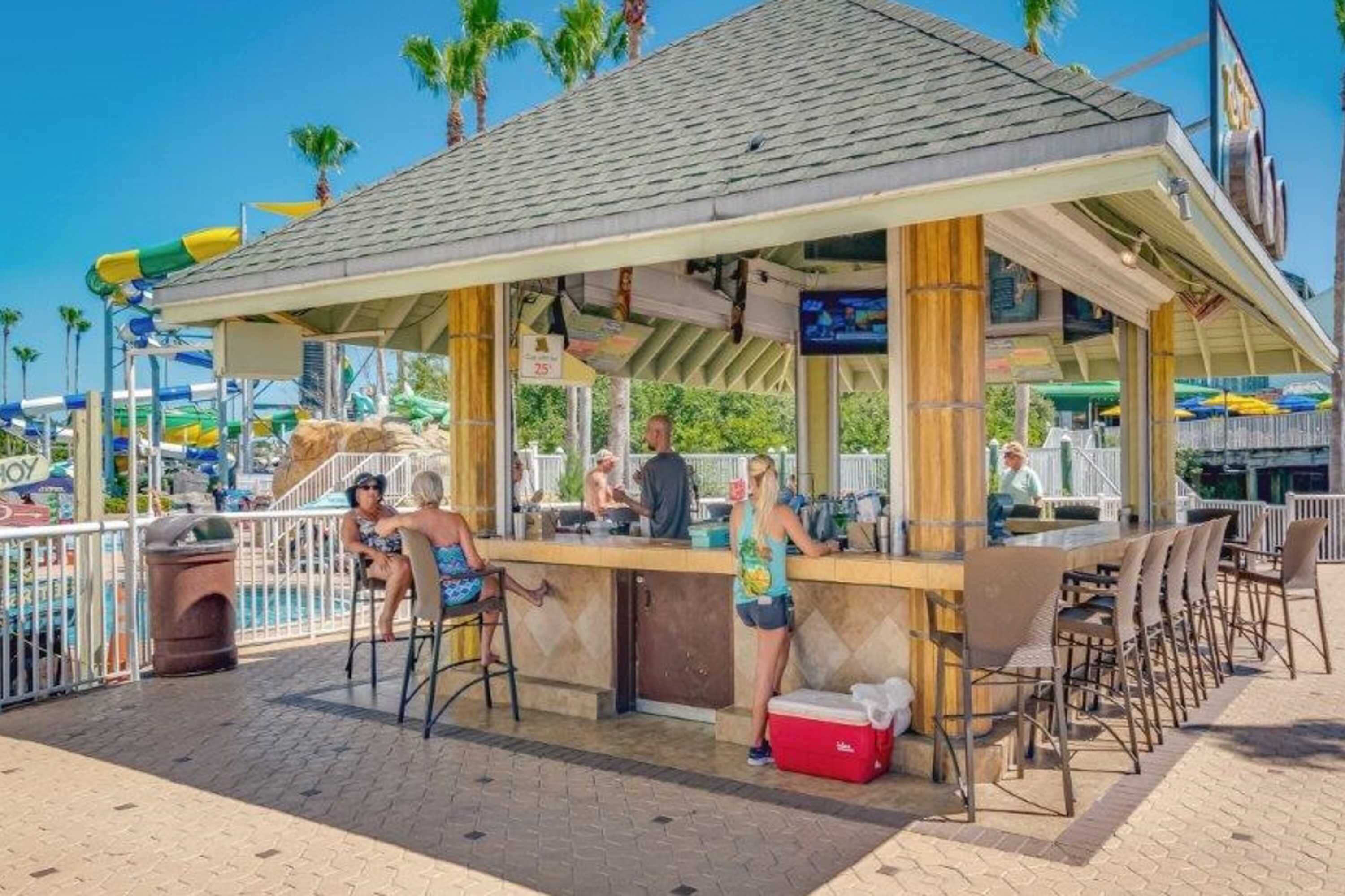 A beachside bar with outdoor seating, a pool in the background, and palm trees.