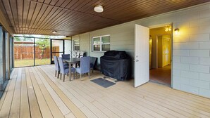 Een overdekt terras met een tafel, stoelen, een barbecue en uitzicht op een achtertuin.