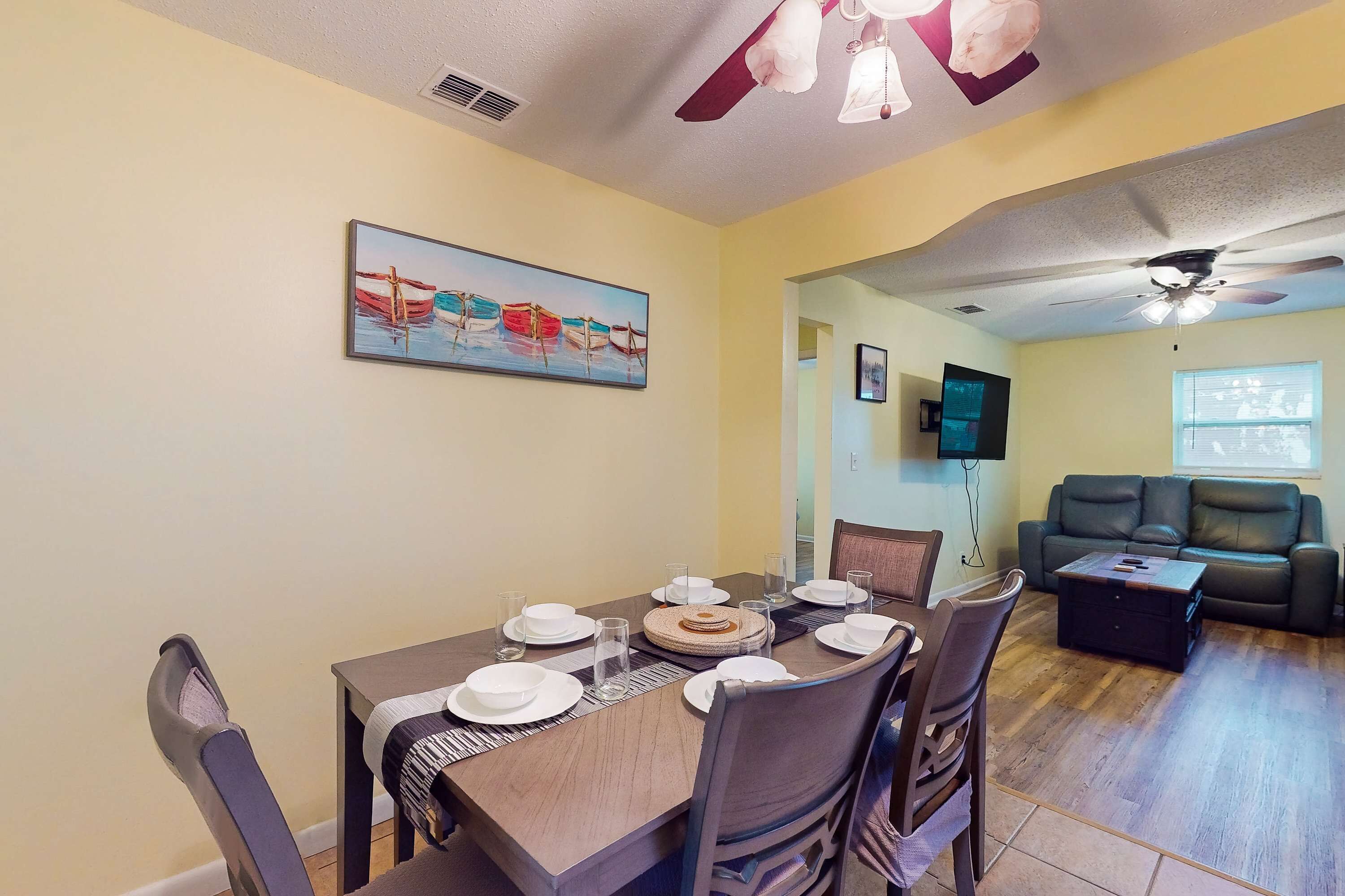A dining area with a wooden table set for four, a ceiling fan, and a painting of boats on the wall.